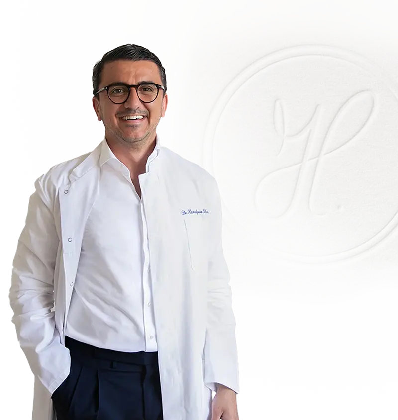 Lächelnder Zahnarzt Dr. Harutyun Oberholzer in weißem Kittel und Brille vor neutralem Hintergrund in seiner Praxis